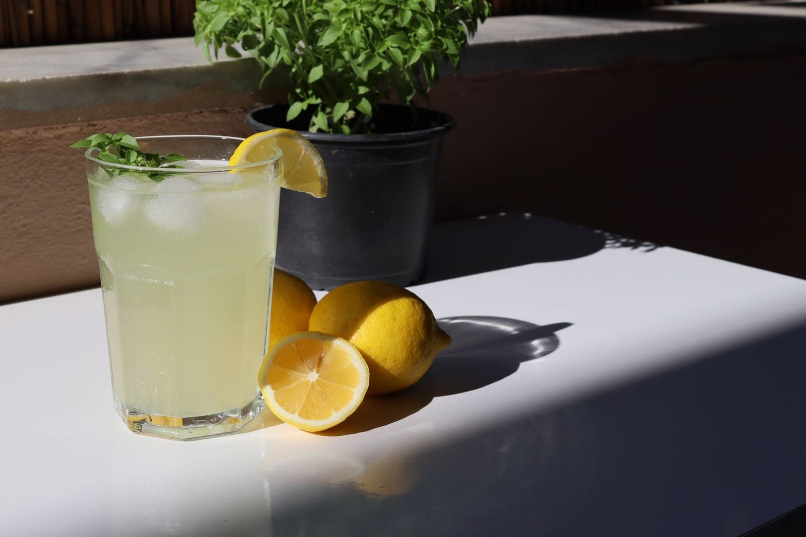 A glass of pale, cloudy lemon barley water with lemons on a sunlit table